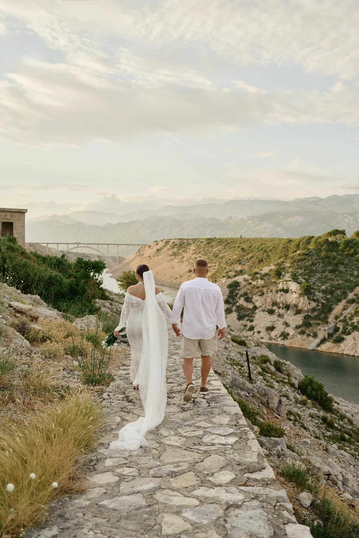 Newlyweds on a coastal stone path with a trailing veil