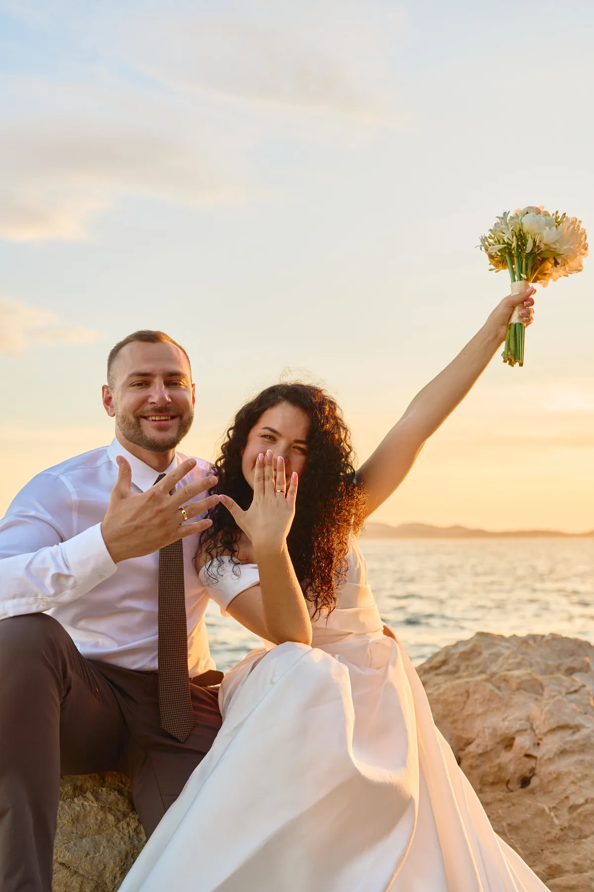Bride laughing and raising her bouquet by the sea